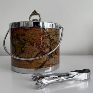 May include: A cylindrical ice bucket featuring a vintage world map print in shades of brown, tan, and cream. The bucket has a polished silver-tone metal rim, base, handle, and a decorative lid knob. A pair of matching silver-tone metal ice tongs is included.