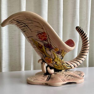 Peut inclure: Un vase en céramique vintage en forme de corne d'abondance, de couleur rose pâle, orné de motifs floraux orange, jaune et violet. Le vase présente une forme incurvée en forme de corne avec des rayures noires sur l'extrémité recourbée.