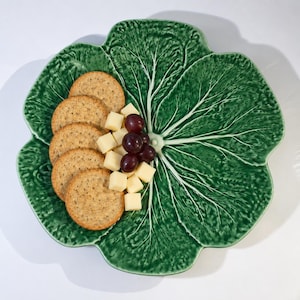 Peut inclure: Une assiette de service en céramique verte en forme de grande feuille de chou, présentant une surface texturée avec des nervures blanches marquées. L'assiette est garnie d'une pile de cinq biscuits ronds, de cubes de fromage et de raisins violets.