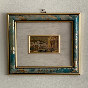 Peut inclure: Une peinture de paysage encadrée représentant un village côtier avec de petits bateaux sur le rivage. L'œuvre est placée dans un passe-partout blanc cassé texturé et un cadre décoratif avec une bordure intérieure dorée et une bordure extérieure marbrée bleue et dorée.