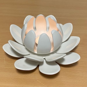 Peut inclure: Un bougeoir en forme de fleur de lotus blanche avec plusieurs pétales superposés. Le centre du bougeoir présente une lumière chaude provenant d'une petite bougie, illuminant l'intérieur des pétales blancs avec un éclat doux aux tons orangés.