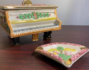 Puede incluir: Una caja decorativa de cerámica con forma de piano con tapa, teclado blanco, detalles dorados y patas negras. Un cenicero en forma de abanico con una escena pintada de figuras y árboles está en primer plano.