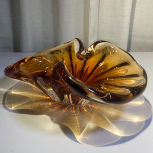 May include: A translucent amber-coloured glass bowl with a wide, organic, undulating rim. The bowl features a deep, flared shape with thick, flowing glass walls that transition from a dark brown base to a lighter, golden-yellow hue at the edges.