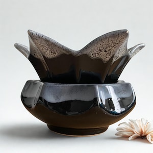 Peut inclure: Un chauffe-cire en céramique composé de deux pièces, arborant une glaçure noire profonde avec un motif de gouttes blanches tachetées sur les bords festonnés. La partie supérieure est un bol aux courbes pétaloïdes sur une base arrondie.