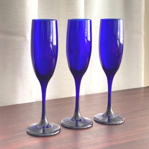 Peut inclure: Trois flûtes à champagne hautes et élancées en verre bleu cobalt vibrant. Chaque verre présente un bol lisse et allongé qui se rétrécit en un pied fin et délicat, reposant sur une base circulaire large pour assurer stabilité et équilibre.