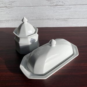 Peut inclure: Un beurrier en céramique blanche avec un couvercle et un petit sucrier avec couvercle. Les deux pièces ont un design géométrique et sont placées sur une surface en bois foncé. Le beurrier est rectangulaire et le sucrier est hexagonal.