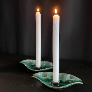 Peut inclure: Deux bougies blanches allumées dans des bougeoirs en céramique vert d'eau en forme de feuille. Les bougeoirs ont une finition brillante et sont placés sur une surface sombre. Les flammes sont vives et le fond est noir.