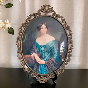 Peut inclure: Portrait ovale d'une femme en robe turquoise, encadré dans un cadre orné de couleur bronze. Le cadre présente un motif floral et de volutes détaillé. Le portrait est exposé sur un support noir.