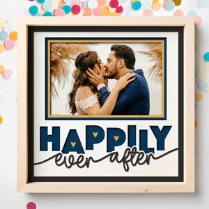 May include: A square wooden frame containing a photograph of a couple kissing, set against a white background. Below the photo, the text "HAPPIILY ever after" is displayed in dark blue and black, featuring small gold heart accents.