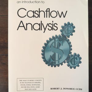 May include: A softcover book titled An Introduction to Cashflow Analysis by Robert J. Donohue CCIM. The white cover features black text and a graphic of three interlocking blue gears, with the largest gear displaying a dollar sign symbol.
