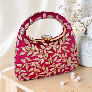 May include: A burgundy handbag with a gold handle and a sparkling gem clasp. The bag is embroidered with gold and silver leaf patterns. The bag is sitting on a wooden surface.