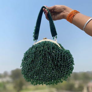 May include: A round, beaded green handbag with a velvet handle. The bag is covered in dangling green beads, creating a fringe effect. The handle is dark green, and the clasp is gold-toned. The bag is held against a blue sky.