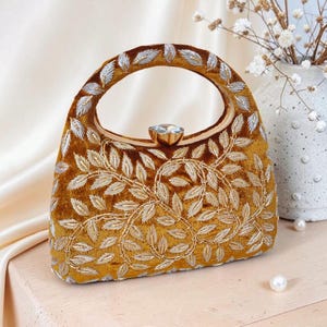 Könnte beinhalten: Eine senffarbene Samt-Handtasche mit ovalem Griff und einem funkelnden Edelsteinverschluss. Die Tasche ist mit aufwendiger Silber- und Goldblattstickerei verziert, die ein luxuriöses und elegantes Design schafft. Ein stilvolles Accessoire.