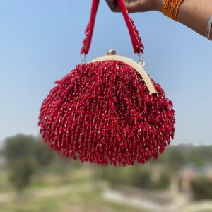 May include: A red beaded handbag with a rounded shape and a gold-colored clasp. The bag has a red handle and is held up against a blue sky. The beads create a textured, fringed effect.