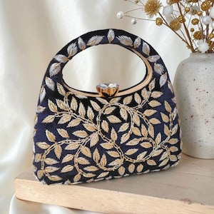 May include: A navy blue handbag with gold leaf embroidery and a gold handle. The bag features a sparkling gemstone clasp. The handle is a gold-toned metal. The bag is sitting on a wooden surface.