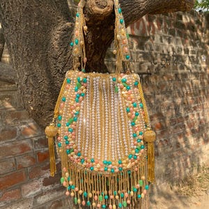 Puede incluir: Un bolso adornado con cuentas, con un marco y asa dorados. El bolso está adornado con cuentas blancas, rosas y turquesas, dispuestas en un patrón floral. Largas borlas doradas cuelgan de la parte inferior. El bolso está colgado de una rama.
