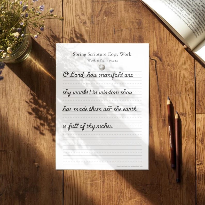 May include: A white sheet of paper with the text "Spring Scripture Copy Work" and a Bible verse written in cursive. Two brown pencils lie next to the paper on a wooden surface. A small vase of flowers is in the upper left corner.