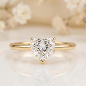 May include: A gold ring featuring a single heart-shaped clear gemstone set in a prong setting. The band is smooth and polished, crafted from yellow gold. The gemstone is faceted, reflecting light with a brilliant white sparkle and clarity.