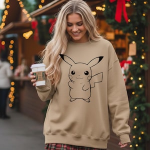 May include: A tan sweatshirt featuring a black outline of Pikachu. The sweatshirt has long sleeves and a crew neck. The person in the image is holding a white coffee cup with a brown sleeve. The background includes Christmas decorations.