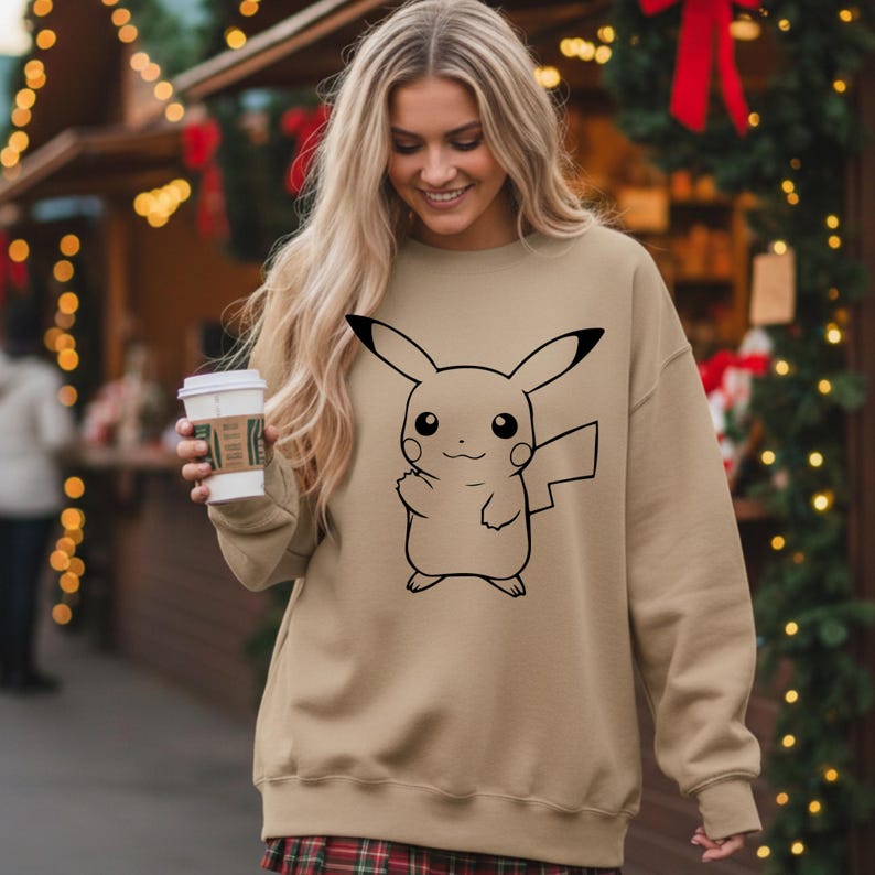 May include: A tan sweatshirt featuring a black outline of Pikachu. The sweatshirt has long sleeves and a crew neck. The person in the image is holding a white coffee cup with a brown sleeve. The background includes Christmas decorations.