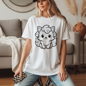 May include: White t-shirt featuring a black and white cartoon fox design. The fox has large eyes, a small nose, and curly hair. The shirt is a short-sleeved crew neck.
