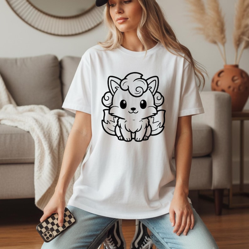 May include: White t-shirt featuring a black and white cartoon fox design. The fox has large eyes, a small nose, and curly hair. The shirt is a short-sleeved crew neck.