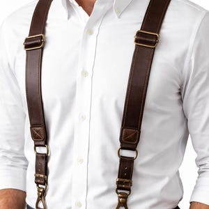 Peut inclure: Bretelles en cuir marron avec des attaches en laiton. Les bretelles sont fixées à une ceinture en cuir marron assortie. La personne porte une chemise blanche boutonnée et un jean bleu. Un accessoire classique.