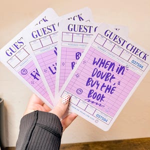 May include: Several light purple and white guest check cards with the words "GUEST CHECK" at the top. The cards have the text "WHEN IN DOUBT, BUY THE BOOK" and other text. The cards are held in a hand.