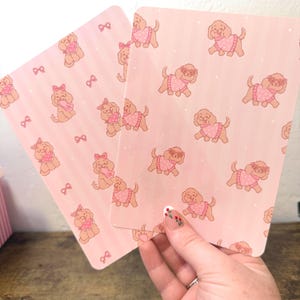 Op de afbeelding: Twee roze rechthoekige kaarten met een herhaald patroon van cartoonhonden met roze truien en strikken. De achtergrond is roze met witte stippen en strikken. Eén kaart heeft verticale strepen.
