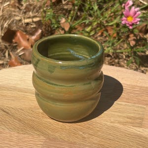 May include: A small, cylindrical ceramic cup with a glossy, olive-green glaze. The exterior features three distinct horizontal ridges or indentations circling the body, creating a textured, undulating surface profile from the base to the rim.