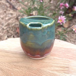 May include: A small, rounded ceramic vessel with a glossy glaze featuring a gradient of deep forest green at the top, transitioning into olive, and ending in a warm reddish-brown base. The rim includes a distinct, integrated horizontal opening.