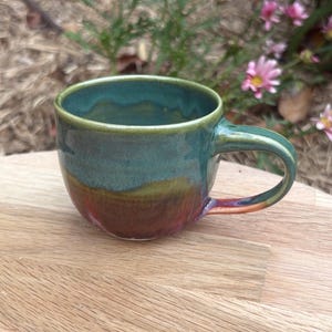 May include: A ceramic mug featuring a rounded body and a curved handle. The exterior and interior glaze transitions from a deep teal at the rim to a warm golden-brown and reddish-purple hue towards the base, creating a smooth, glossy finish.