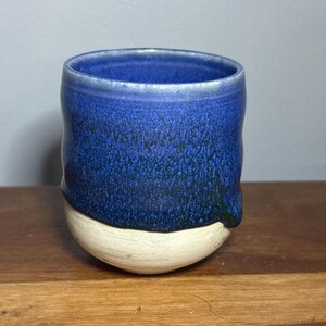 May include: A ceramic cup featuring a two-tone design with a deep blue glazed upper section and a matte off-white base. The blue glaze has a textured, speckled appearance that drips slightly over the unglazed, light-coloured bottom portion.