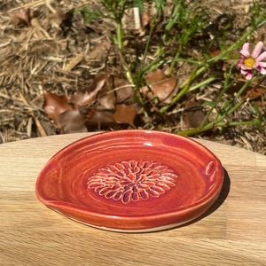 May include: A round ceramic spoon rest with a glossy red and orange glaze. The centre of the dish features a raised, textured floral pattern resembling a dahlia or chrysanthemum, with a smooth, slightly curved rim for holding kitchen utensils.