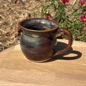 May include: A round ceramic mug featuring a glossy glaze with blended tones of deep mahogany brown and muted sage green. The mug has a wide, curved body, a flared rim, and a sturdy, arched handle attached to the side, with a white base.