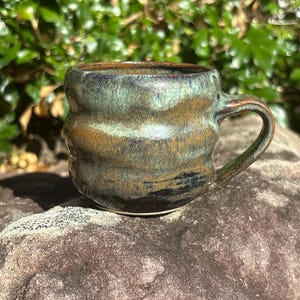 May include: A handcrafted ceramic mug with a distinctive, textured pattern. The mug has a dark, earthy colour scheme with hues of green, brown, and blue. The handle is curved, and the mug appears to be resting on a rock.
