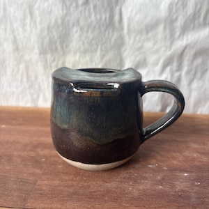 May include: A handcrafted ceramic mug with a distinctive, curved rim. The mug has a dark, glossy glaze with hues of blue, green, and brown. The base is white, and it has a curved handle. The mug is designed for beverages.