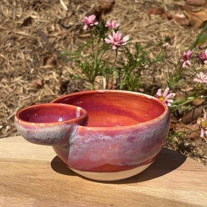May include: A ceramic snack bowl featuring a large main compartment and a smaller attached side compartment. The piece has a glossy glaze with blended shades of vibrant red, deep pink, and soft lavender, transitioning to a plain cream base.
