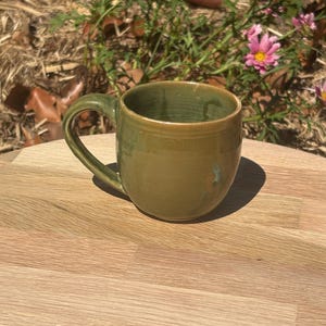 May include: A round ceramic mug with a glossy olive green glaze and a smooth finish. The mug features a curved handle on the side and a wide opening at the top. The exterior surface shows subtle variations in the green colour, reflecting light.