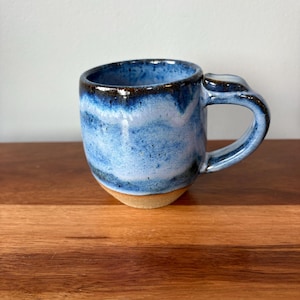 May include: A ceramic mug featuring a rounded body with a glossy glaze in shades of deep blue, light blue, and white. The rim is dark, and the base is unglazed, showing a natural tan clay colour. It includes a sturdy, curved handle on the side.