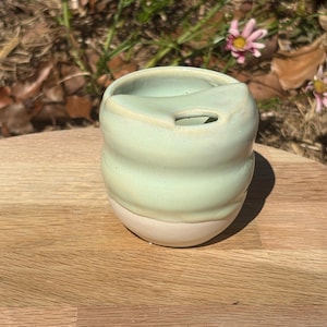 May include: A small, rounded ceramic cup featuring a smooth, pale mint green glaze on the upper portion and an unglazed, matte off-white base. The rim has a distinct, moulded indentation on one side, creating a unique shape for the vessel.