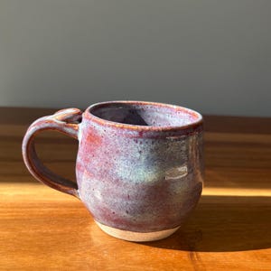 May include: A ceramic mug with a rounded body and a curved handle. The exterior features a mottled glaze with blended tones of deep purple, reddish-brown, and hints of soft blue. The base of the mug is unglazed, revealing a light beige clay texture.