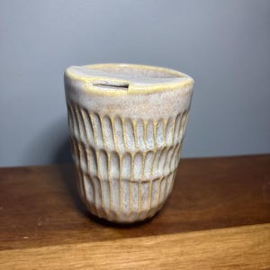 May include: A ceramic travel mug with a lid, featuring a textured exterior with vertical carved grooves. The mug has a light beige and cream glaze with yellow-toned highlights along the rim and the textured ridges of the body.