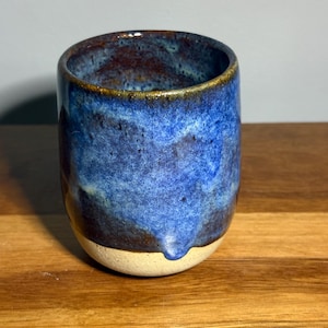 May include: A ceramic cup with a rounded shape, featuring a glossy glaze in shades of deep blue and dark brown on the upper portion, transitioning to a speckled, matte off-white finish on the lower base, with a visible drip of blue glaze.