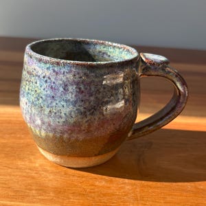 May include: A ceramic mug with a rounded body and a curved handle. The exterior features a speckled glaze in shades of blue, purple, and green, transitioning to a solid brown base. The rim is dark, and the handle includes a small thumb rest.