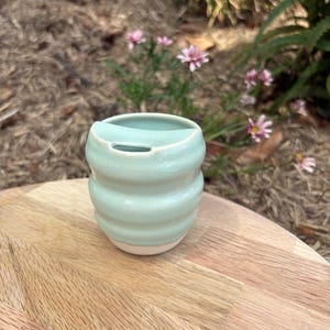 May include: A light mint green ceramic cup with a smooth, glossy finish and three horizontal ridges along the body. The cup features a unique, integrated lid design with a small, narrow opening at the top rim, showcasing a simple, functional form.