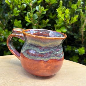 May include: A ceramic mug with a rounded body and a curved handle. The glaze features a gradient of deep reddish-brown at the base, transitioning into a mottled band of sage green and blue, with a smooth, glossy finish on the rim and handle.