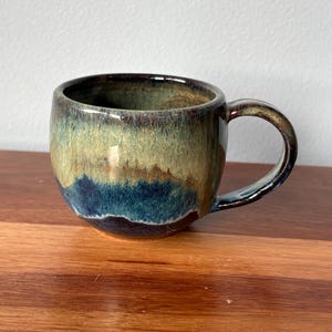 May include: A ceramic mug with a rounded body and a curved handle. The mug has a glaze with a gradient of colours, including blue, green, and brown. The mug is on a wooden surface.