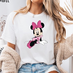 May include: White t-shirt featuring a Minnie Mouse graphic. The cartoon character is depicted in a pink polka-dot dress and bow. The word "WHITE" is printed in black at the top left corner of the shirt.