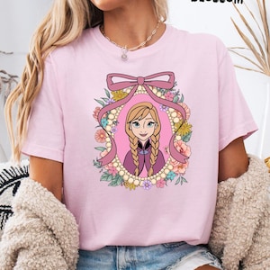 May include: A light pink t-shirt featuring a cartoon illustration of a woman with blonde braids, framed by a pink bow and floral elements. The word "BLOSSOM" is printed in black at the top. The shirt is a casual wear item.